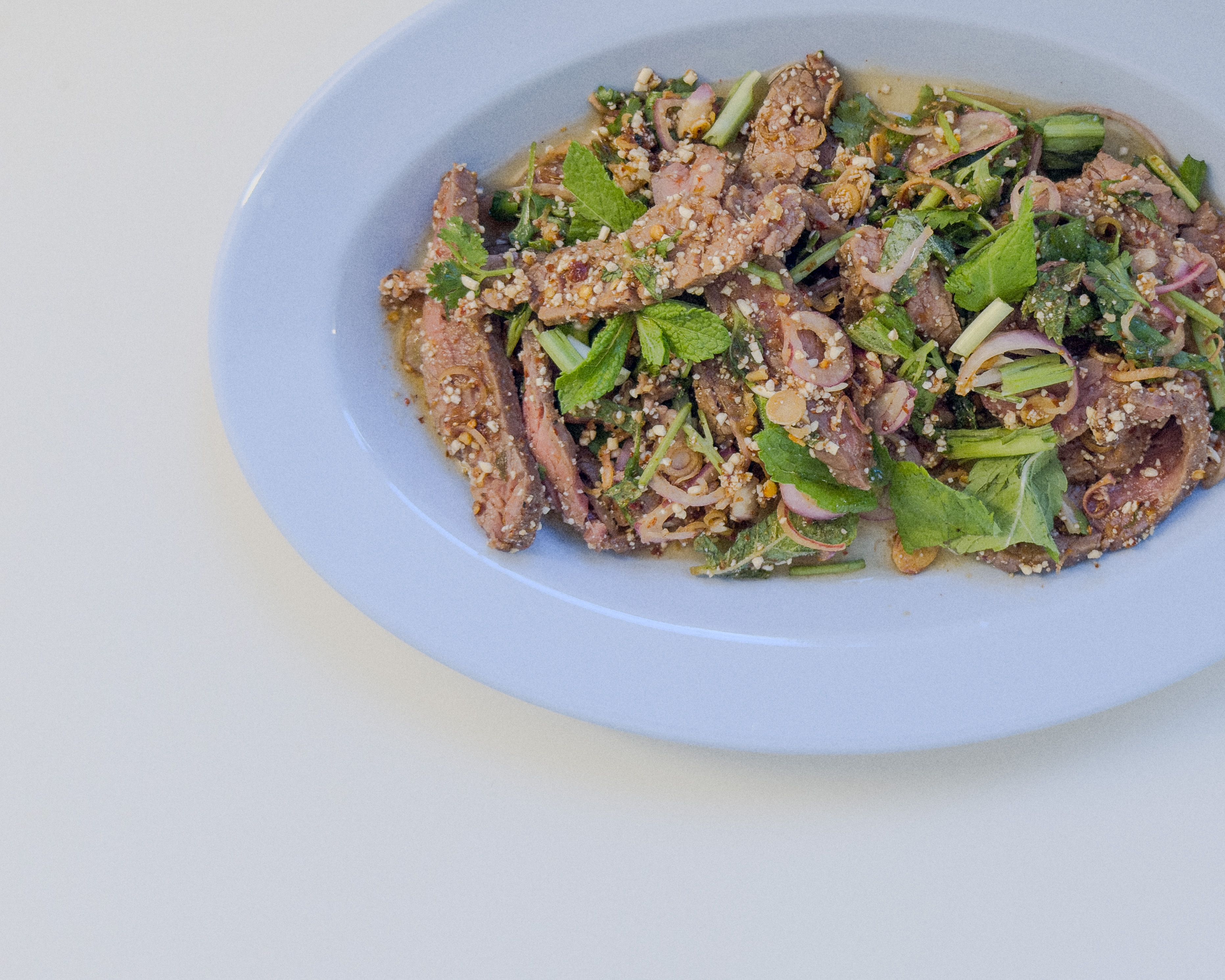 Authentisches Nam Tok Nuea – gegrillter Rindfleischsalat mit Minze, Schalotten, Chili und geröstetem Reispulver auf blauem Teller