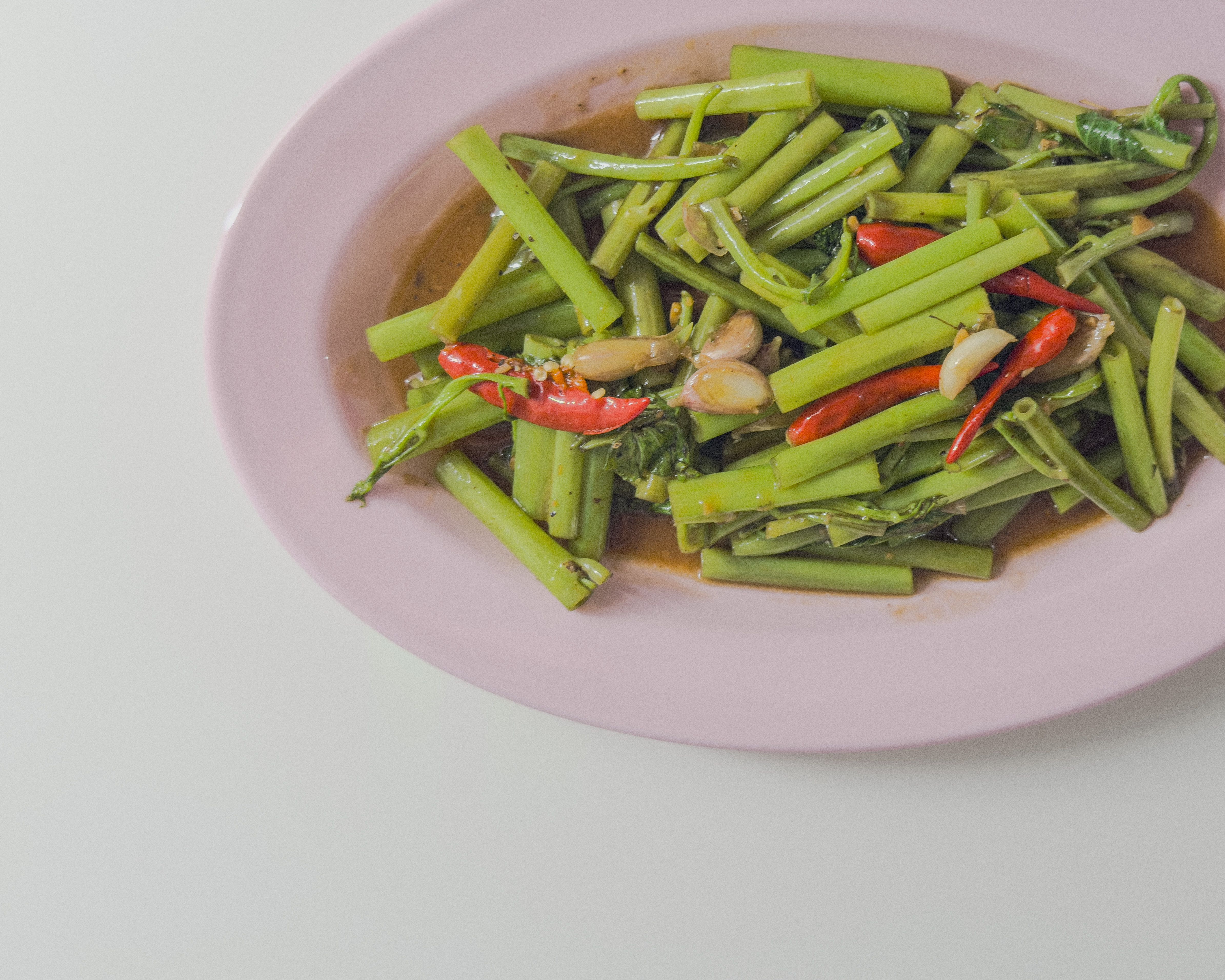 Authentisches Pak Boong Fai Daeng – thailändischer gebratener Wasserspinat mit Knoblauch, Chili und Sauce auf pinkem Teller.
